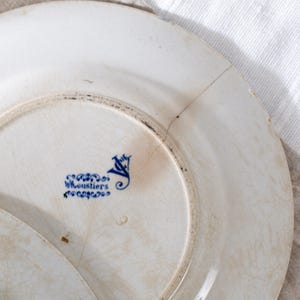 May include: Close-up of a white antique plate with a blue floral design and the text "W.Loustiers". The plate has a cracked surface and a dark rim. The plate is part of a vintage dishware set.
