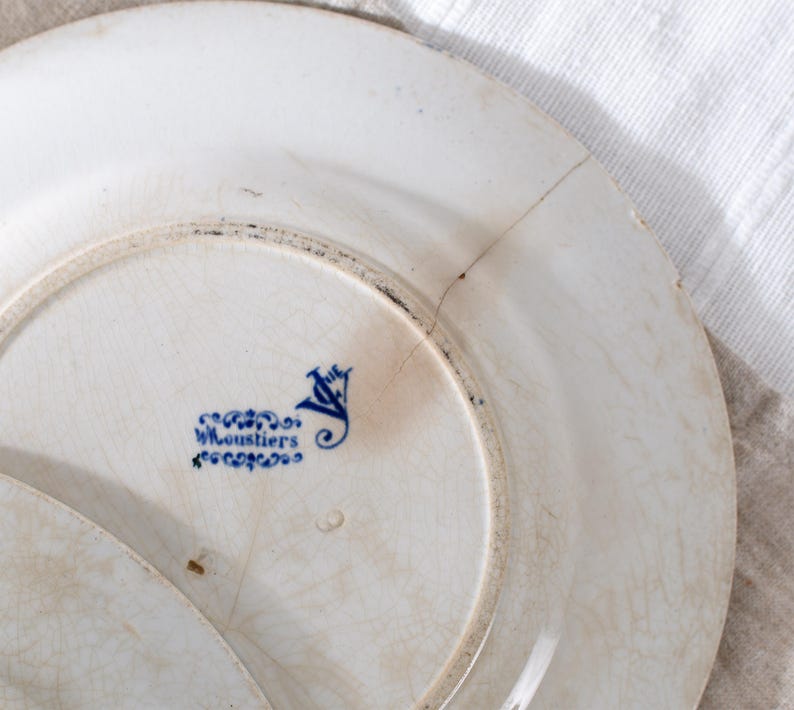 May include: Close-up of a white antique plate with a blue floral design and the text "W.Loustiers". The plate has a cracked surface and a dark rim. The plate is part of a vintage dishware set.