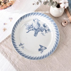 May include: White ceramic plate with a blue bird and floral design. The plate features a central illustration of a bird perched on a flowering branch, with additional bird and floral motifs around the rim. The plate has a decorative blue border.