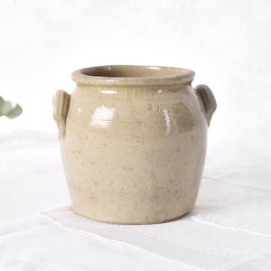 Könnte beinhalten: Ein beigefarbener, antiker Steinguttopf mit zwei kleinen Henkeln. Der Topf hat einen runden Körper und einen leicht ausgestellten Rand. Die Oberfläche weist feine horizontale Linien auf und eine verblasste "2 N"-Markierung. Der Topf steht auf einer weißen, strukturierten Oberfläche.