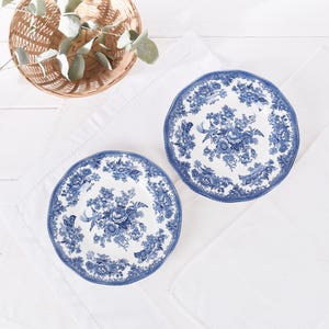 May include: Two white ceramic plates with a blue floral design. The plates have a scalloped edge and are placed on a white cloth. A woven basket with greenery is in the background.