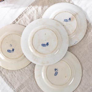 May include: Four antique white ceramic plates with blue floral designs on the underside. The plates have visible signs of wear, including cracks and discoloration, suggesting age and use. The plates are arranged on a neutral-colored linen cloth.