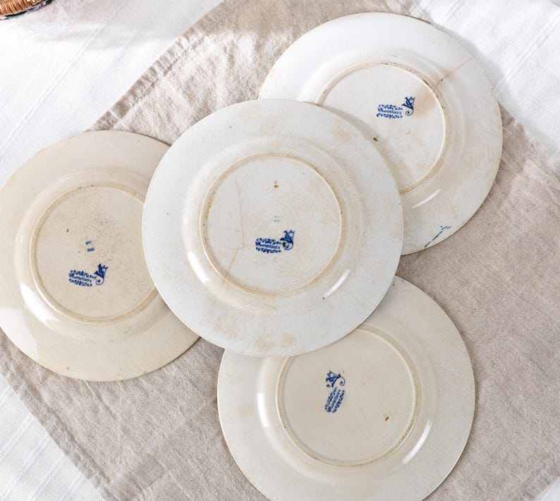 May include: Four antique white ceramic plates with blue floral designs on the underside. The plates have visible signs of wear, including cracks and discoloration, suggesting age and use. The plates are arranged on a neutral-colored linen cloth.