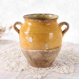May include: A two-handled earthenware pot featuring a glossy mustard yellow glazed upper section and a raw, unglazed brown clay base. The vessel has a wide, rounded body with visible surface wear, chips in the glaze, and a thick, uneven rim.