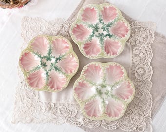 May include: Three ceramic oyster plates featuring a scalloped, shell-like design. Each plate has six individual compartments with a soft pink finish, separated by intricate, raised green foliage patterns that radiate from the center.
