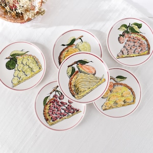 Peut inclure: Un ensemble de six assiettes rondes en céramique blanche, chacune ornée d'une illustration peinte à la main d'une part de tarte aux fruits. Les assiettes présentent un fin liseré rouge et illustrent diverses garnitures aux fruits.