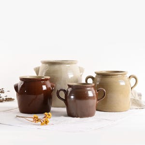 Könnte beinhalten: Vier Keramikkrüge mit zwei braunen und zwei beigefarbenen glasierten Oberflächen. Die Krüge haben jeweils zwei Henkel und sind auf einer weißen Oberfläche angeordnet.
