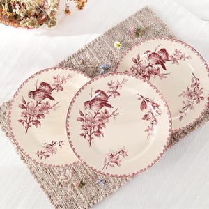 May include: Three round ceramic plates with a cream-colored base and a dark red transferware pattern. Each plate features a detailed illustration of birds perched on flowering branches, with a matching floral border around the outer rim.