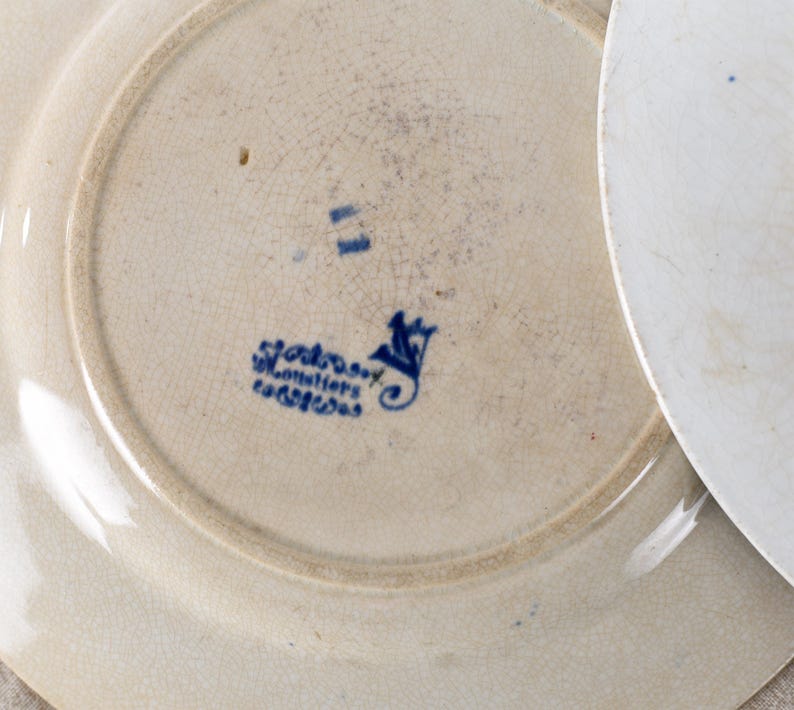 May include: Close-up of two antique white ceramic plates. The plates have a cream-colored base with a blue floral design and text. The plates have a slightly aged appearance with visible cracks in the glaze, suggesting age and use.