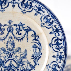 May include: Close-up of a white and blue antique plate. The plate features a detailed blue floral and cherub design around the rim and center. The plate has a slightly aged appearance with visible cracks, adding to its vintage charm.