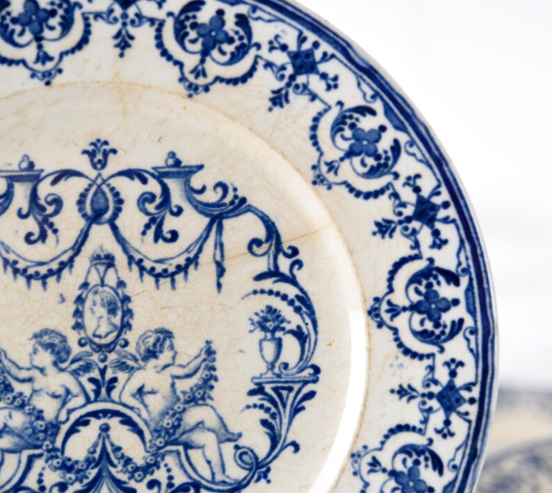 May include: Close-up of a white and blue antique plate. The plate features a detailed blue floral and cherub design around the rim and center. The plate has a slightly aged appearance with visible cracks, adding to its vintage charm.