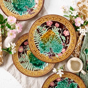 Peut inclure: Un ensemble d'assiettes rondes en céramique présentant un relief de feuilles vertes, de fougères et de fleurs roses sur un fond brun foncé. Chaque assiette possède une large bordure jaune moutarde avec un motif géométrique grec répétitif.