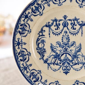 May include: A white ceramic plate with a blue floral and cherub design. The plate features a detailed border and a central motif of two cherubs holding garlands, with a portrait in the center. The design is reminiscent of antique tableware.