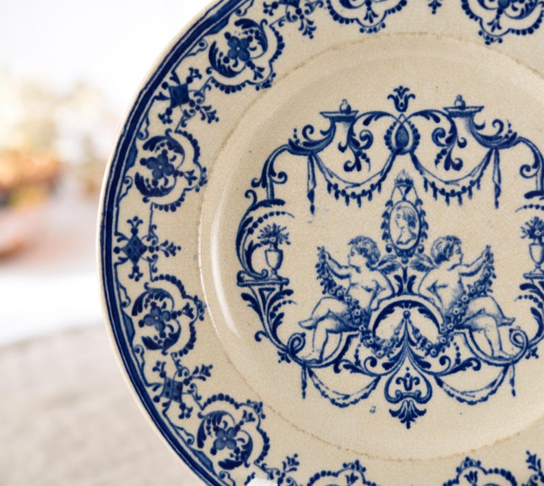 May include: A white ceramic plate with a blue floral and cherub design. The plate features a detailed border and a central motif of two cherubs holding garlands, with a portrait in the center. The design is reminiscent of antique tableware.