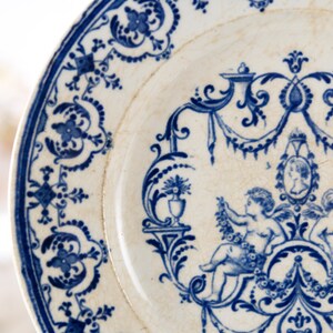 May include: A close-up of a white plate with a blue floral design. The plate features a detailed border and a central design with cherubs and a portrait. The plate has a vintage, antique aesthetic.