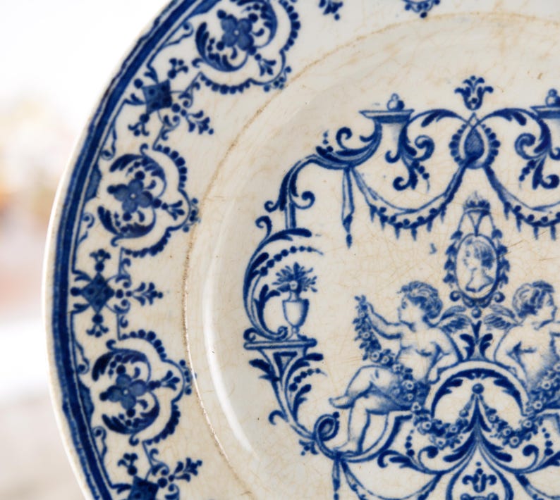 May include: A close-up of a white plate with a blue floral design. The plate features a detailed border and a central design with cherubs and a portrait. The plate has a vintage, antique aesthetic.