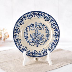 May include: A vintage-style ceramic plate with a cream-colored base and intricate blue floral patterns. The plate features a central design of cherubs and decorative elements, and is displayed on a white stand. The plate's rim is also adorned with blue patterns.