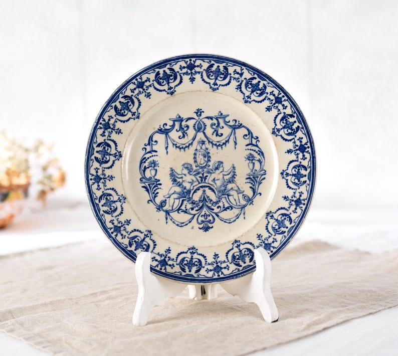 May include: A vintage-style ceramic plate with a cream-colored base and intricate blue floral patterns. The plate features a central design of cherubs and decorative elements, and is displayed on a white stand. The plate's rim is also adorned with blue patterns.