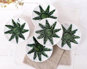 Puede incluir: Conjunto de cinco platos de cerámica blanca con superficie texturizada. Cada plato presenta un círculo central verde oscuro y un diseño de estrella radiante en verde, que se asemeja a una planta estilizada. Los platos están dispuestos sobre una tela de lino blanca y beige.