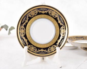 May include: A decorative plate with a black and gold ornate design. The plate has a white center and is displayed on a white stand. The gold detailing features intricate patterns. Additional plates are visible in the background.
