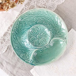 Puede incluir: Un plato redondo de cerámica de color verde claro con un motivo floral central en relieve rodeado de intrincados patrones de hojas. La superficie esmaltada tiene un acabado brillante con sutiles variaciones de color que resaltan el relieve botánico.