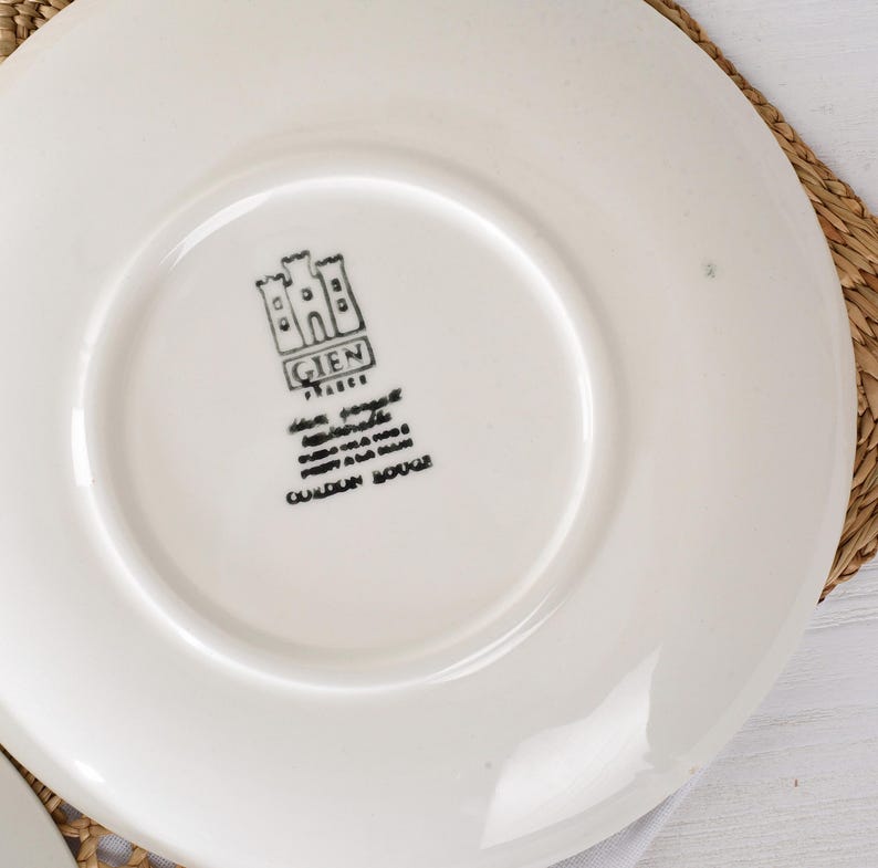 Puede incluir: Plato de cer&aacute;mica blanca con logotipo y texto en negro. El logotipo presenta un dise&ntilde;o de castillo con la palabra "GIEN" encima del texto. El plato est&aacute; sobre una estera tejida.