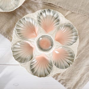May include: A white ceramic oyster plate with six shell-shaped compartments and a central well. The shells are shaded with pink and gray, creating a delicate, elegant design. The plate is placed on a neutral-colored linen surface.