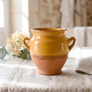 Könnte beinhalten: Ein rustikale Keramikvase mit zwei Henkeln. Der obere Teil ist goldgelb, mit einem Terrakotta-Sockel. Die Vase steht auf einer neutralen Leinentischdecke und erinnert an einen Vintage- oder Landhausstil. Die Vasenhöhe beträgt ca. 20 cm.