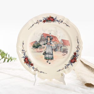 Puede incluir: Un plato de cerámica blanco con un borde elevado y un diseño floral alrededor del borde. El centro del plato presenta una escena pintada de una mujer con un vestido tradicional de pie frente a una granja. La escena está rodeada por un borde de flores rojas y azules.