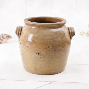 May include: A vintage, tan stoneware crock with two side handles. The antique jar has a wide opening and a slightly textured surface. Horizontal lines encircle the upper portion of the crock, adding a decorative element to the rustic design.