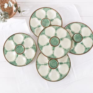 May include: Five ceramic oyster serving platters. Each platter has a green and white seashell design with a central dip bowl. The platters are arranged on a white cloth, with a wicker basket and greenery in the background.