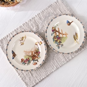 Puede incluir: Dos platos redondos de cerámica con una base de color crema y un borde decorativo con pequeñas flores azules. Cada plato muestra una ilustración colorida de una valla de madera, varias flores de jardín, espigas de trigo y una mariposa.
