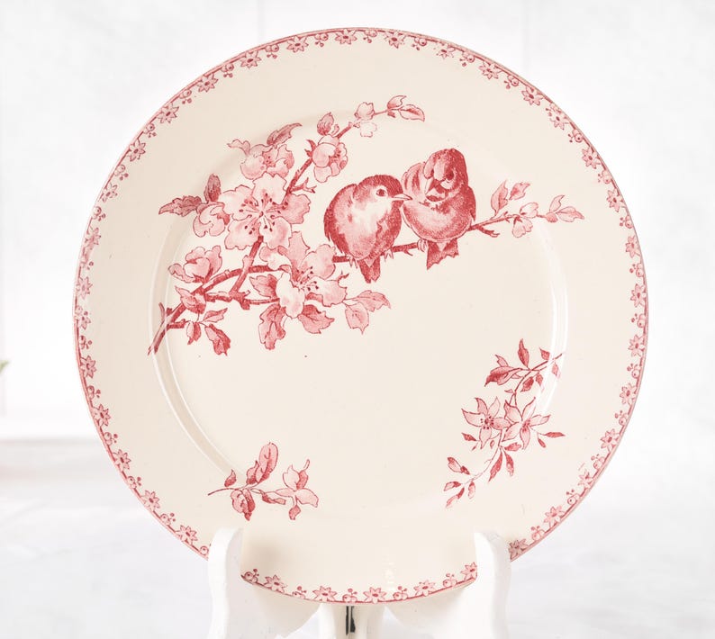 Puede incluir: Plato de cer&aacute;mica color crema con dise&ntilde;o floral y de p&aacute;jaros en rojo. El plato presenta un borde de peque&ntilde;os motivos florales y una ilustraci&oacute;n central de dos p&aacute;jaros posados en una rama florecida.