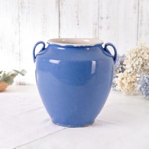 May include: A blue ceramic vase with two handles and a cream-coloured rim. The vase has a glossy finish and a rounded shape. Decorative vase for home decor.