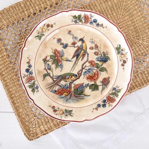 May include: Cream-coloured decorative plate with a scalloped edge and a maroon border. The plate features a detailed illustration of two birds perched on a flowering branch with red, blue, and yellow blossoms. The plate is set on a woven mat.