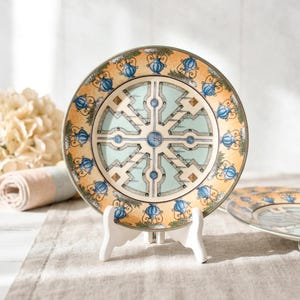 May include: Decorative ceramic plate with a garden design. The plate features a light blue and beige color scheme with a detailed pattern of a garden layout. The plate is displayed on a white stand, with a rolled-up cloth and flowers in the background.