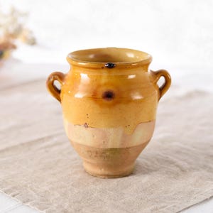 May include: A small, antique, yellow and brown ceramic vase with two handles. The vase has a rounded body and a wide opening. The color is a mix of yellow, brown, and beige, with a rustic, aged appearance. The vase is sitting on a beige linen cloth.