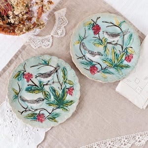 Puede incluir: Dos platos redondos de cerámica con borde festoneado, con fondo azul claro, ilustraciones en relieve de pequeños pájaros grises posados en ramas marrones, hojas verdes y racimos de pequeñas bayas rojas en un patrón natural.