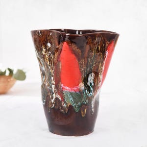 May include: A distinctive ceramic vase with an irregular, organic form. The vase has a deep brown base, transitioning to a striking red and brown upper section. The textured surface is accented with white, green, and gold.