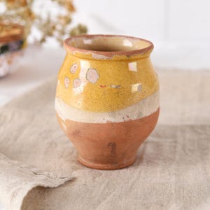Peut inclure: Petit pot en céramique rustique, corps arrondi et large ouverture. Le pot présente un dégradé de jaune, blanc et marron. La partie jaune présente des taches roses délavées. Le pot est probablement fait à la main et a une esthétique vintage.