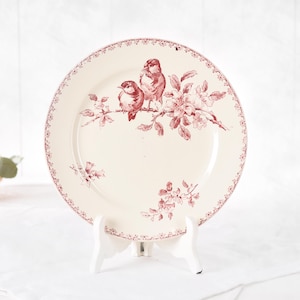 Puede incluir: Un plato de cer&aacute;mica color crema con un dise&ntilde;o de p&aacute;jaros y flores granate. El plato presenta dos p&aacute;jaros posados en una rama con flores. El borde tiene un borde granate decorativo. El plato se exhibe en un soporte blanco.