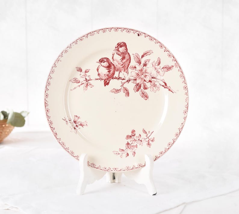 Puede incluir: Un plato de cer&aacute;mica color crema con un dise&ntilde;o de p&aacute;jaros y flores granate. El plato presenta dos p&aacute;jaros posados en una rama con flores. El borde tiene un borde granate decorativo. El plato se exhibe en un soporte blanco.