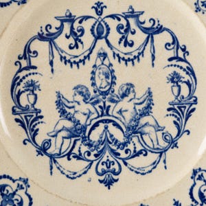 May include: Close-up of a white ceramic plate with a blue floral and cherub design. The central motif features two cherubs holding garlands, framed by ornate blue scrollwork and a portrait. The plate's edge has a repeating floral pattern.