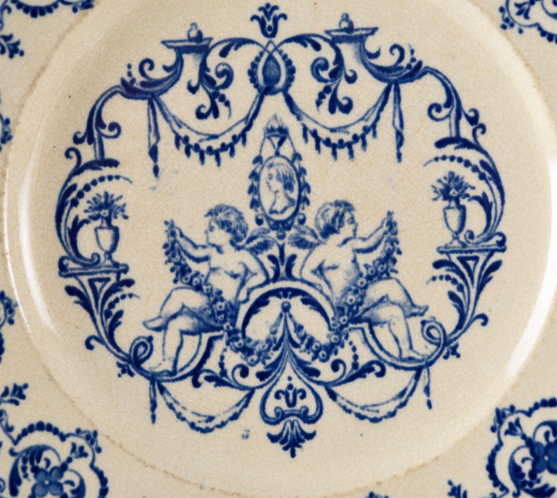May include: Close-up of a white ceramic plate with a blue floral and cherub design. The central motif features two cherubs holding garlands, framed by ornate blue scrollwork and a portrait. The plate's edge has a repeating floral pattern.