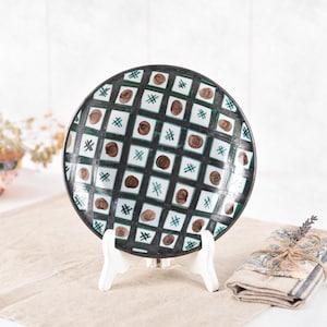 May include: A decorative ceramic plate with a black rim, featuring a grid design of white squares, brown circles, and green crosshatch details. The plate is presented on a white stand, with a folded cloth and dried flowers visible in the background.