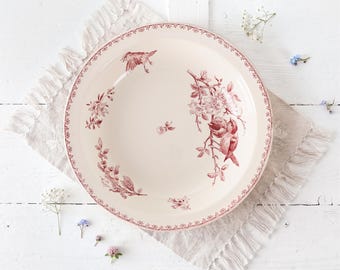 May include: A round ceramic plate with a cream-colored base and a deep red transferware pattern. The design features detailed illustrations of birds perched on flowering branches, a small central floral motif, and a decorative floral border.