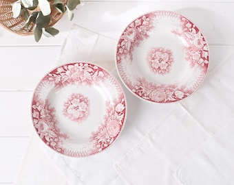 May include: Two white ceramic plates with a red floral pattern. The plates have a central floral design and a border of flowers and leaves. The plates are on a white cloth.