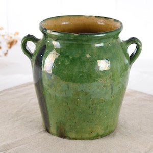 May include: A green ceramic pot with two handles. The pot has a wide opening and a slightly textured surface. The interior is a light brown color, and the exterior is a mottled green with some darker areas. The pot is sitting on a neutral-colored cloth.