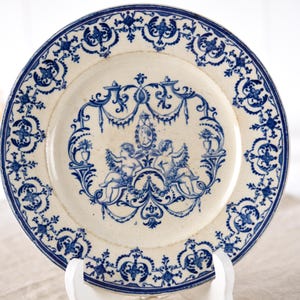 May include: Antique white and blue decorative plate. The plate features a detailed blue floral border and a central design with cherubs, vases, and ornate scrollwork. A vintage piece for display or dining.