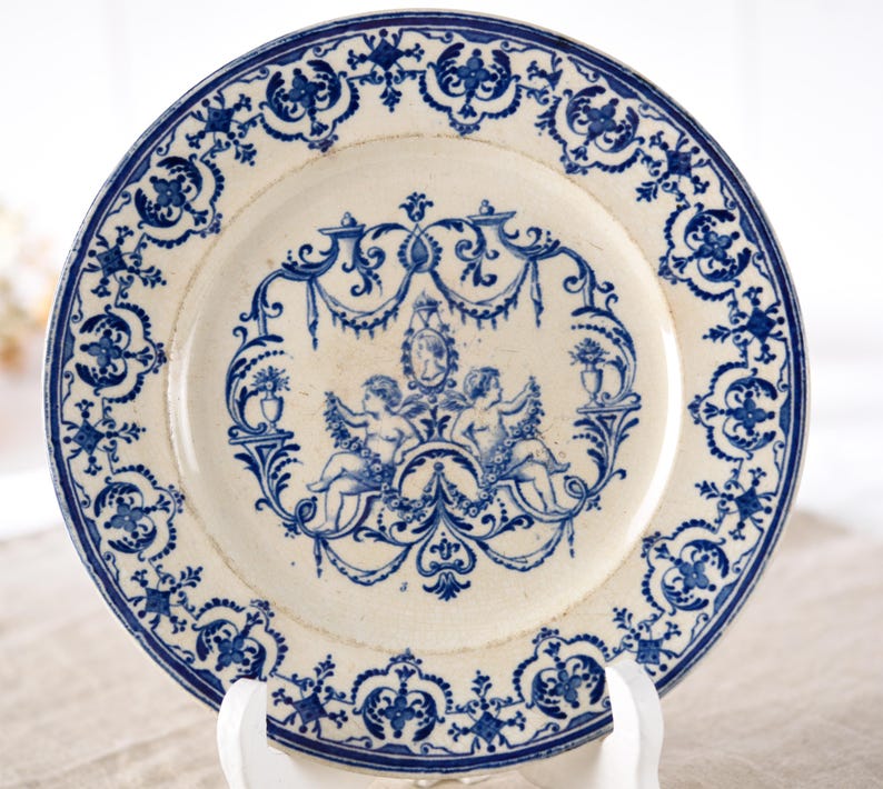 May include: Antique white and blue decorative plate. The plate features a detailed blue floral border and a central design with cherubs, vases, and ornate scrollwork. A vintage piece for display or dining.